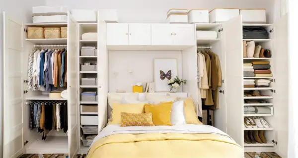 Habitación matrimonio pequeña con armario blanco, cama con ropa de cama amarilla y almohadas, y estanterías llenas de ropa y cestas.